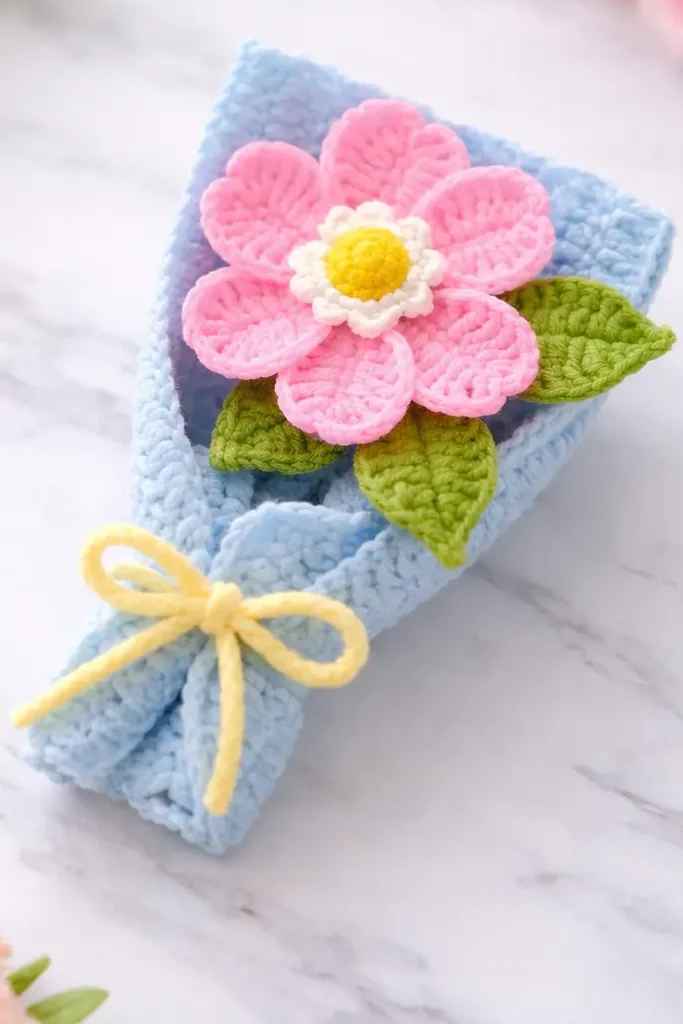 Crochet mini flower bouquet on a marble background with a blue wrapper, pink flower, and yellow bow